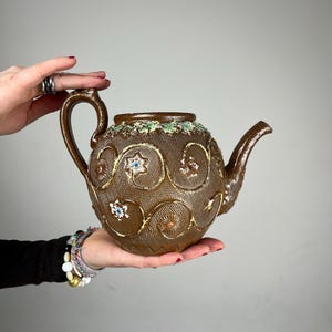 Könnte beinhalten: Braune Keramik-Teekanne mit strukturierter Oberfläche und floralen und wirbelnden Designs. Die Teekanne hat einen Griff und einen Ausguss und ist mit farbenfrohen Akzenten verziert. Ein einzigartiges Stück für Teeliebhaber.