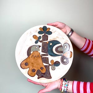 May include: A white ceramic plate with a brown, blue, and green stylized design of two birds on a tree branch. The plate is hand-painted and has a rustic, vintage look.