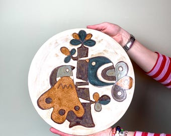 May include: A white ceramic plate with a brown, blue, and green stylized design of two birds on a tree branch. The plate is hand-painted and has a rustic, vintage look.