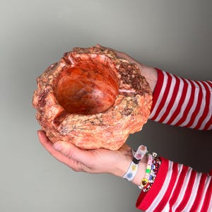 Peut inclure: Un bol en pierre sculptée, orange-rouge, avec un intérieur arrondi. Le bol a un extérieur rugueux et texturé et est tenu dans deux mains. Les mains portent des bracelets et la personne porte une chemise à manches longues rayée rouge et blanche.