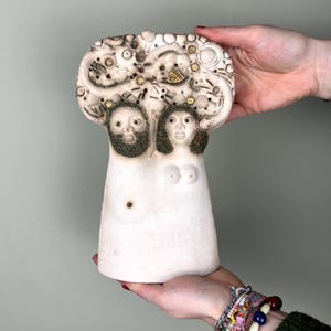 Może przedstawiać: Biała rzeźba ceramiczna przedstawiająca dwie stylizowane twarze z teksturowanymi włosami i brodami. Rzeźba ma pękatą górę z osadzonymi elementami dekoracyjnymi i jest trzymana przez dwie ręce. Rzeźba ma około 25 cm wysokości.