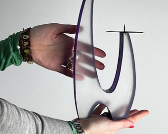 May include: Two clear, abstract candle holders with a purple accent. The candle holders have a modern, curved design with a metal disc at the top to hold the candles. The holders are being held up by a person's hands.