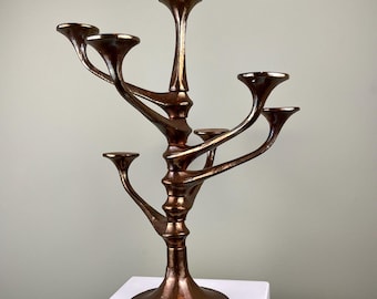 May include: A copper-toned metal candelabra with five arms, resembling a stylized tree branch. The candelabra has a wide base and a smooth, polished finish.