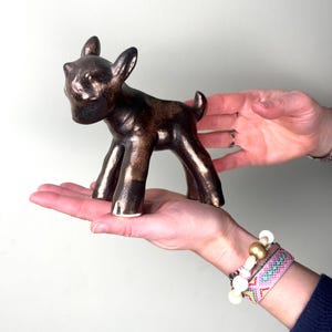 Könnte beinhalten: Bronze farbene Keramik-Hirschfigur mit glänzendem Finish. Der Hirsch hat eine stilisierte Form mit großen Ohren und einem kleinen Schwanz. Die Figur wird in den Händen einer Person gehalten.