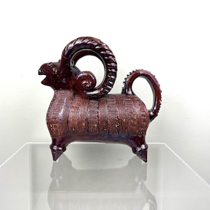 Può includere: Brocca in ceramica a forma di ariete, in una ricca tonalità marrone scuro. Il vaso presenta un design dettagliato con motivi strutturati e un manico curvo. Le corna dell'ariete sono ben visibili.