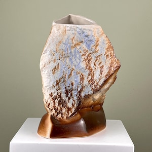 Könnte beinhalten: Eine Keramikvase in Form eines Steins mit brauner und weißer Glasur und blauen Akzenten. Die Vase hat eine raue, strukturierte Oberfläche.