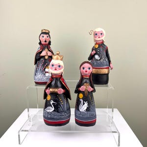 Pode incluir: Conjunto de quatro figuras de cerâmica pintadas à mão que representam os três reis magos e um pastor. As figuras estão vestidas com túnicas pretas e douradas com detalhes brancos. Estão colocadas sobre um expositor de acrílico de dois níveis.