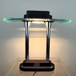 Könnte beinhalten: Eine moderne Schreibtischlampe mit einem schwarzen rechteckigen Oberteil und Sockel, getragen von zwei zylindrischen Pfosten in Chromfarbe. Ein horizontales, gebogenes, transparentes grünes Glasregal erstreckt sich von oben. Die Lampe ist an eine Steckdose angeschlossen.