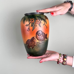 May include: A vintage ceramic vase with a brown, green, and orange glaze. The vase features a painted design of two birds perched on a branch with flowers.