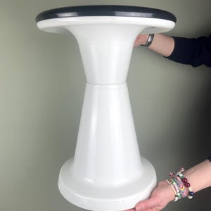 Può includere: Uno sgabello bianco a forma di clessidra con piano nero. Lo sgabello ha una base ampia e circolare e un piano svasato. Il design è semplice e moderno, adatto a vari stili di interni. Lo sgabello è tenuto da due mani.