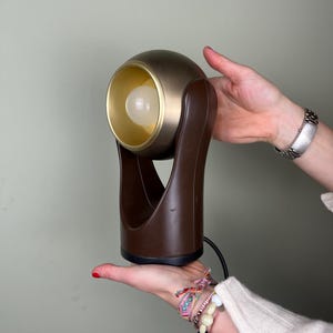 May include: A vintage brown and gold accent lamp with a spherical head and a brown base. The lamp features a gold-colored interior and a visible lightbulb. The lamp is held by a person.