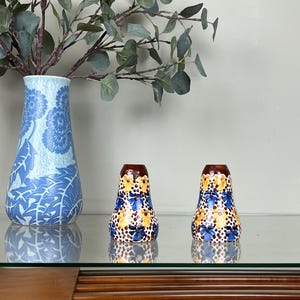 Könnte beinhalten: Zwei dekorative Salz- und Pfefferstreuer mit floralem Design. Die Streuer sind weiß mit blauen und gelben Blumen und braunen Punkten. Die Oberteile sind braun. Eine blaue Vase mit floralem Design und ein Eukalyptuszweig befinden sich im Hintergrund.