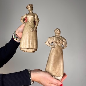 Op de afbeelding: Twee beige, mat afgewerkte beeldjes van figuren in lange jurken met pofmouwen. Eén figuur draagt een hoed en houdt een klein voorwerp vast, terwijl de andere de handen in de zij heeft en het hoofd omhoog gericht houdt met een gladde textuur.