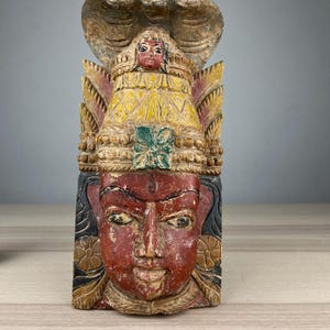 May include: A carved wooden sculpture of a deity with a red face, yellow clothing, and a cobra hood. The sculpture is painted in various colours and shows signs of age and wear, suggesting it is an antique or vintage piece.
