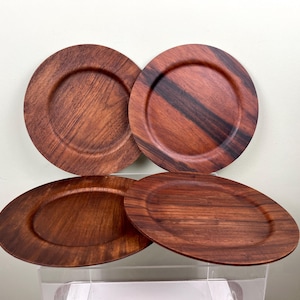 Può includere: Quattro piatti in legno di colore marrone scuro con venature visibili. Due sono rotondi e due ovali. I piatti hanno un bordo leggermente rialzato e una finitura liscia e lucida, perfetti per servire il cibo.