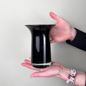 May include: A black glass vase with a wide mouth and a narrow base. The vase is being held in a person's hands.