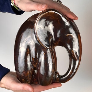 May include: A brown ceramic elephant figurine with a glossy finish. The elephant is in a sitting position with its trunk curled up.