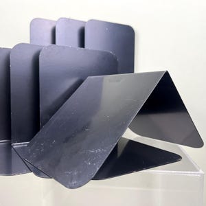 May include: A set of black metal bookends with a modern design. The bookends are made of folded metal sheets, creating a series of angled supports. The bookends are designed to hold books upright on a shelf or desk.