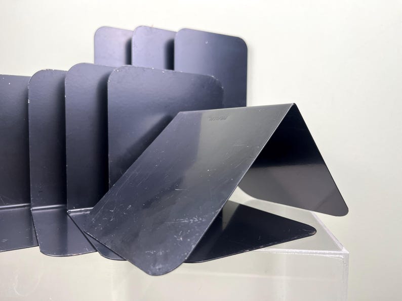 May include: A set of black metal bookends with a modern design. The bookends are made of folded metal sheets, creating a series of angled supports. The bookends are designed to hold books upright on a shelf or desk.