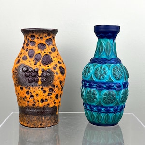 Könnte beinhalten: Zwei Keramikvasen. Eine ist orange mit dunkelbraunen Flecken und einer strukturierten Oberfläche. Die andere ist blau und türkis mit einem Blumenmuster. Beide Vasen haben ein einzigartiges, dekoratives Design.