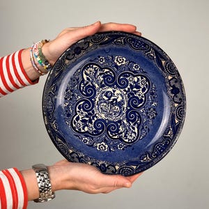 Könnte beinhalten: Ein runder Keramikteller in tiefblauer und weißer Farbgebung. Das Design umfasst komplizierte Blumenmuster, geschwungene Ornamente und ein zentrales Emblem, das eine Löwenfigur zeigt, umgeben von dekorativen Rändern und detaillierten Motiven.