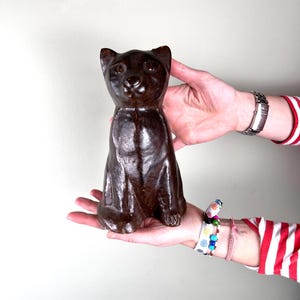 Może przedstawiać: Ceramiczna figurka kota w kolorze ciemnobrązowym w pozycji siedzącej. Figurka ma szczegółową twarz z małym nosem i dużymi oczami.