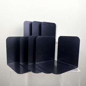 May include: A collection of six black metal bookends. Each bookend features a right-angled design with a curved top. The bookends are positioned upright, in a staggered arrangement, ideal for organizing books on a shelf or desk.