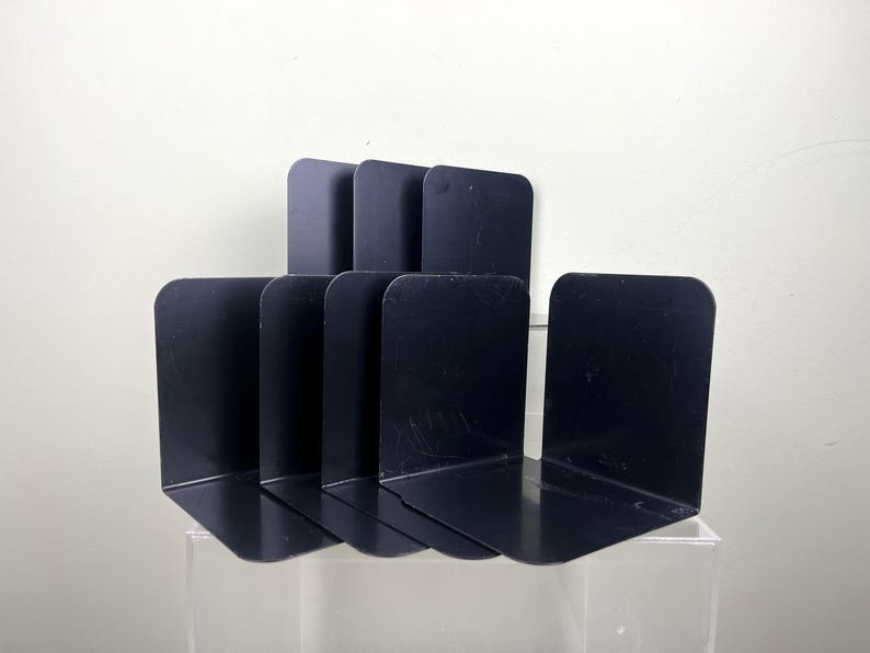May include: A collection of six black metal bookends. Each bookend features a right-angled design with a curved top. The bookends are positioned upright, in a staggered arrangement, ideal for organizing books on a shelf or desk.