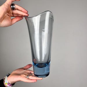 Op de afbeelding: Een hoge, doorschijnende glazen vaas met een gladde, golvende rand en een taps toelopend silhouet. Het glas verloopt van een diep, verzadigd blauw bij de zware voet naar een lichtere, rookgrijze tint aan de bovenkant.