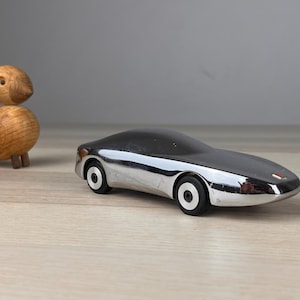 Può includere: Un'auto giocattolo in cromo lucido con una carrozzeria aerodinamica e liscia, dotata di pneumatici neri con cerchi bianchi e un piccolo emblema quadrato rosso sul cofano anteriore, accanto a una piccola statuetta di uccello in legno chiaro.