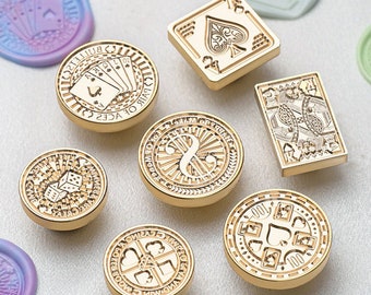 May include: A set of nine gold-toned metal wax seals with various designs, including playing cards, dice, and a musical note. The seals are arranged on a white surface.