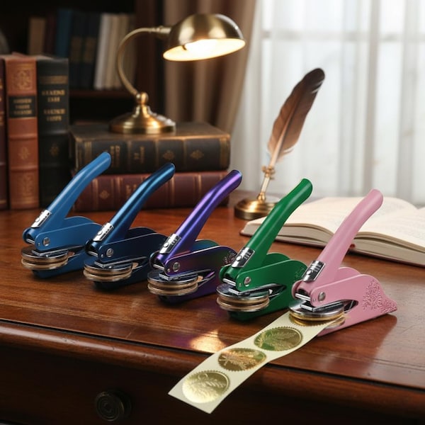 May include: A selection of embossing seals in various colours: blue, navy, purple, green, and pink. Each seal features a lever handle and a circular die. A strip of gold foil seals is in the foreground, with a desk lamp and books in the background.