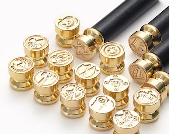 May include: A collection of gold-colored wax seal stamps with various embossed designs, including stars, flowers, and geometric shapes. Each stamp has a round head and a short, cylindrical base. The stamps are paired with black wooden handles.