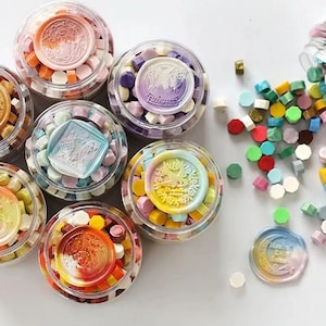K&ouml;nnte beinhalten: Sortiment an bunten Wachssiegel-Stempeln in durchsichtigen Gl&auml;sern mit verschiedenen Designs. Jedes Glas enth&auml;lt eine Vielzahl von Wachssiegel-Perlen in verschiedenen Farben und Formen. Die Wachssiegel sind perfekt, um Briefen, Einladungen und anderen Papierarbeiten einen Hauch von Eleganz zu verleihen.