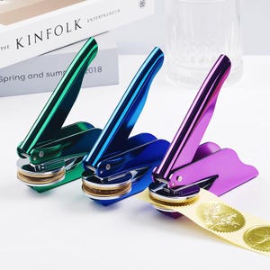 Könnte beinhalten: Drei metallische Siegelpressen in Grün, Blau und Lila. Jede Presse hat einen langen Griff und eine kreisförmige Basis. Ein Streifen goldener Folienaufkleber wird von der lila Presse gestempelt. Das Buch "The Kinfolk Entrepreneur" ist im Hintergrund.