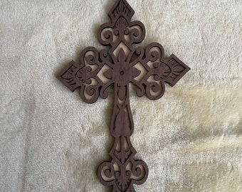 May include: A dark brown wooden cross with an ornate, openwork design. The cross features intricate patterns and scrollwork, with a lighter-colored backing visible through the cutouts. The cross is a decorative religious item.