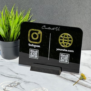 May include: A black acrylic contact information display with gold-colored Instagram and website icons. The display includes QR codes and the text "Contact Us" in a script font. It is supported by a black base and sits on a marble surface.