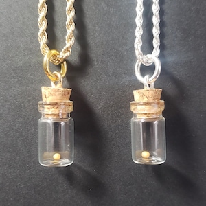 Könnte beinhalten: Zwei Halsketten mit Glasfläschchen, die jeweils eine einzelne Perle enthalten. Die Fläschchen sind mit Korkstopfen verschlossen. Eine Halskette hat eine goldene Kette, die andere eine silberne Kette.
