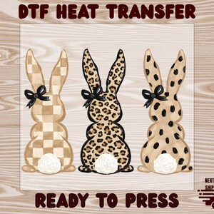 May include: DTF heat transfer featuring three Easter bunny designs. The bunnies have different patterns: checkered, leopard print, and spotted. Each bunny has a black bow and a white fluffy tail. The words "READY TO PRESS" are at the bottom.