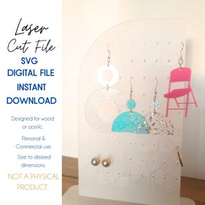 May include: A white display stand with holes for hanging earrings. There are three pairs of earrings hanging on the stand, including a pair of turquoise and white earrings, a pair of clear and white earrings, and a pair of pink chair earrings. The text "Laser Cut File SVG DIGITAL FILE INSTANT DOWNLOAD" is printed on the left side of the image.