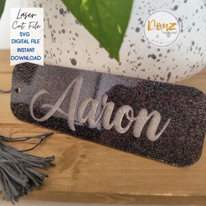 May include: A rectangular, glittery black name tag with the name "Aaron" in a silver script font. The tag has a small hole at the top with a gray tassel attached. Text on the left side reads "Laser Cut File SVG Digital File Instant Download".