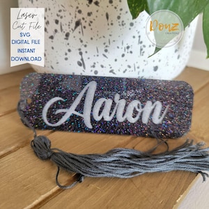 May include: A rectangular, glittery name tag with the name "Aaron" in white script. The tag is dark with iridescent glitter and has a gray tassel attached. Text on the left reads "Laser Cut File SVG Digital File Instant Download."
