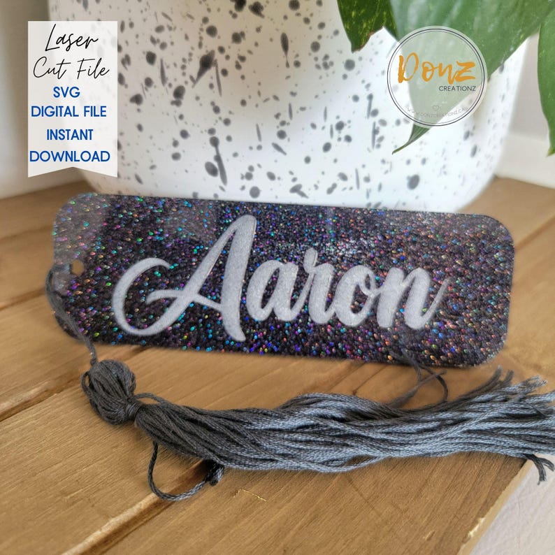 May include: A rectangular, glittery name tag with the name "Aaron" in white script. The tag is dark with iridescent glitter and has a gray tassel attached. Text on the left reads "Laser Cut File SVG Digital File Instant Download."
