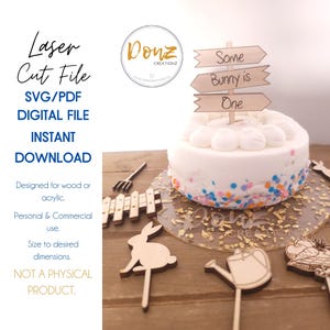 May include: A set of laser cut wooden cake toppers for a first birthday party. The toppers include a bunny, a watering can, a fence, a rake, a basket of eggs, and a sign that says "Some Bunny is One".