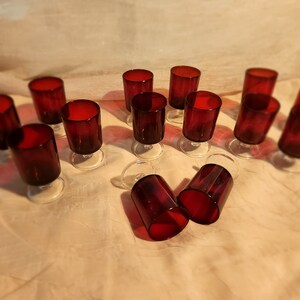 May include: A set of ruby red wine glasses with clear stems. The glasses have a cylindrical shape with a rounded base. The image shows the glasses arranged on a light-coloured surface, with some upright and others tipped over.