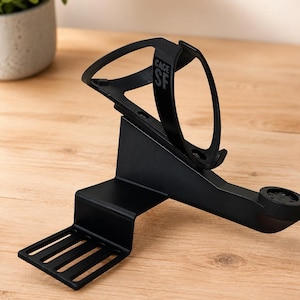 May include: A matte black bicycle water bottle cage with an integrated computer mount. The cage features the text CAGE SF printed on the side. The structure includes a flat, slotted mounting base and a curved arm extending to a circular mount.