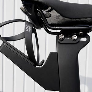 Könnte beinhalten: Schwarzer Fahrrad-Flaschenhalter an einem schwarzen Fahrradsattel befestigt. Der Käfig ist mit "CAGE SF" beschriftet. Der Sattel ist schwarz und trägt den Markennamen "fizik". Die Befestigungshardware ist sichtbar.