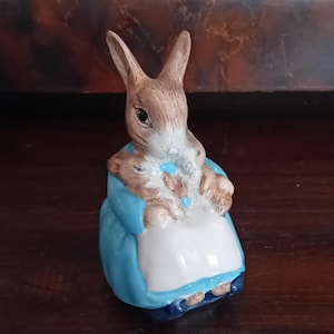 May include: A ceramic figurine of a brown rabbit wearing a blue jacket and a white apron. The rabbit is holding a smaller brown rabbit in its arms. The figurine features painted details including black eyes, whiskers, and blue buttons on the jacket.