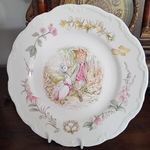 Peut inclure: Une assiette en céramique blanche à bord festonné ornée d'une illustration centrale représentant un renard et un canard vêtus de vêtements. Le bord est décoré d'éléments botaniques peints, dont des fleurs roses et jaunes, un nid d'oiseau et deux petits canetons jaunes.