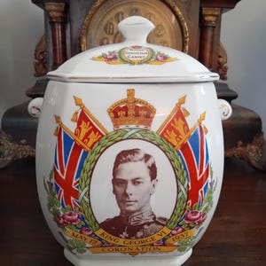 Peut inclure: Un pot en céramique blanche avec couvercle, orné d'un portrait du roi George VI, d'une couronne et de drapeaux Union Jack. Le texte indique Royal Souvenir Casket, King George VI, May 12 1937 et Coronation. Il possède deux anses latérales.